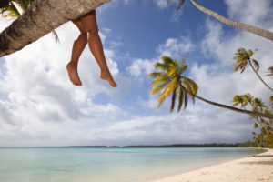 girl sitting on a palm tree dangling her feet over the sandy beaches of Long Island with aqua waters
